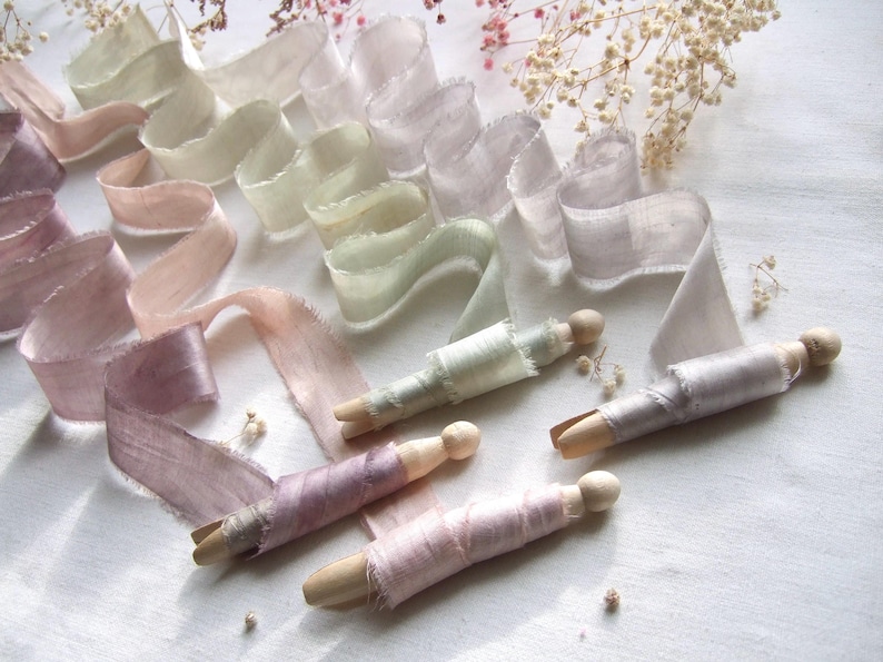 May include: A collection of frayed-edge silk ribbons in soft pastel shades of mauve, dusty pink, sage green, and light gray. Several ribbons are neatly wound around wooden clothespins, while others are draped in loose, flowing waves.