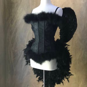 May include: A black corset with a fur trim and a black feather boa. The corset is on a white mannequin with black angel wings.