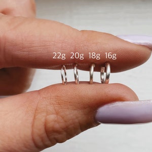May include: Four silver hoop earrings in different sizes, labelled 22g, 20g, 18g, and 16g. The hoops are held in a person's hand.
