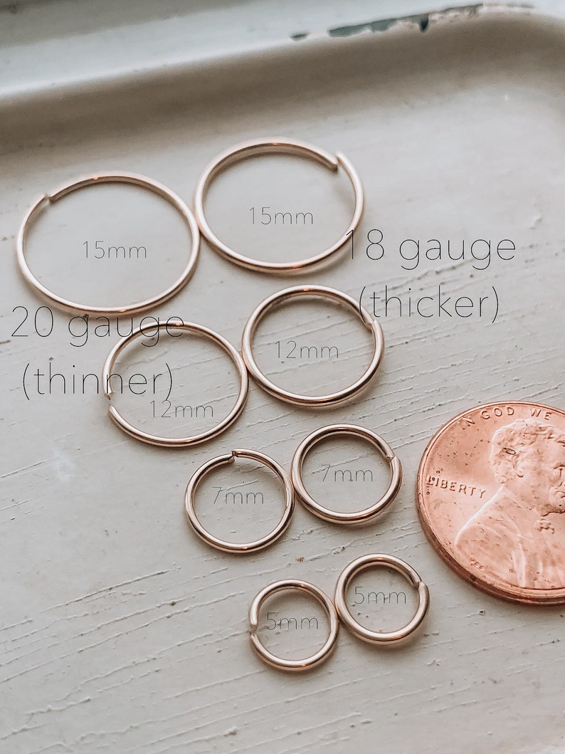 May include: A set of gold hoop earrings in various sizes, ranging from 5mm to 15mm. The earrings are arranged in a circle with a penny for scale. The text "18 gauge (thicker)" and "20 gauge (thinner)" is visible in the image.