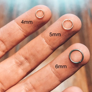 May include: Three gold nose rings in different sizes, 4mm, 5mm, and 6mm, are held by a hand. The rings are shown on the fingers of the hand.