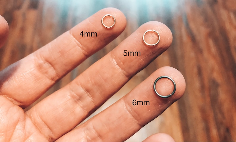 May include: Three gold nose rings in different sizes, 4mm, 5mm, and 6mm, are held by a hand. The rings are shown on the fingers of the hand.