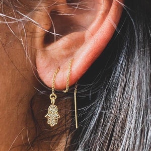 May include: A gold chain earring with a hamsa hand charm and a thin gold bar charm. The charms are dangling from the earlobe.