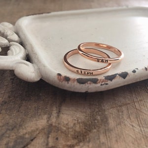 May include: Two gold-toned rings with the names "Steph" and "Sam" engraved on them. The rings are on a white ceramic dish with a decorative handle.