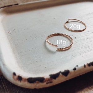 May include: Two gold rings on a white tray. The rings are both twisted and have a beaded texture. The larger ring is labelled "20g" and the smaller ring is labelled "18g".