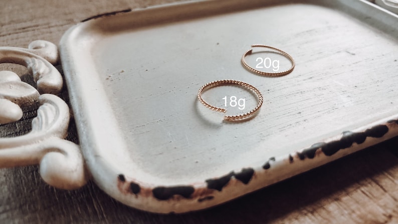 May include: Two gold rings on a white tray. The rings are both twisted and have a beaded texture. The larger ring is labelled "20g" and the smaller ring is labelled "18g".