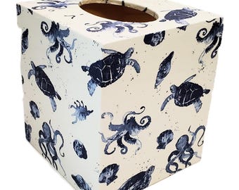 May include: A square tissue box with a cream-colored background. The box is decorated with navy blue sea turtles, octopuses, and seashells. The top has an oval opening for tissue dispensing.