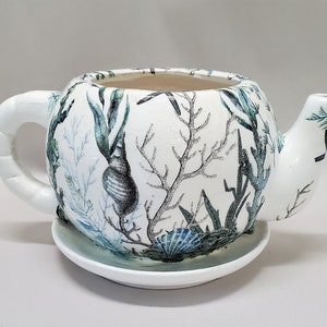 May include: White ceramic teapot with a seashell and seaweed design. The teapot has a round body, a curved handle, and a spout. It sits on a matching saucer. The design features blue, green, and gray elements.