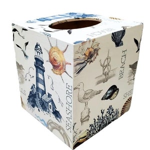 May include: Square tissue box with a coastal theme. The box is white with illustrations of a lighthouse, seashells, seagulls, and nautical knots. The words "Seashore", "Beach", and "Shells" are printed on the sides.