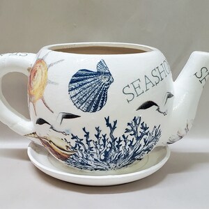 May include: White ceramic teapot with seashell and coral illustrations. The teapot features a handle, spout, and a saucer base. The word "SEASHORE" is printed on the side. Decorative coastal themed item.