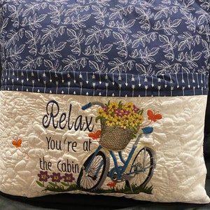 May include: A decorative pillow with a navy blue leaf pattern on top and a white quilted bottom. The bottom features an embroidered bicycle with a basket of flowers and the text "Relax You're at the Cabin".