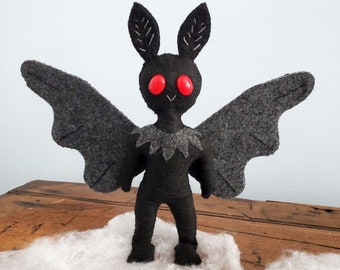May include: A black felt bat with red button eyes. The bat has large wings and is standing on a wooden surface with white spider webbing.