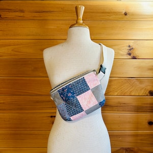 Op de afbeelding: Een patchwork heuptas met een crèmekleurige riem en zwarte gesp wordt op een mannequin getoond. De tas heeft stoffen in marineblauw, roze en grijs, met bloemen- en geometrische patronen. De tas heeft een ritssluiting.