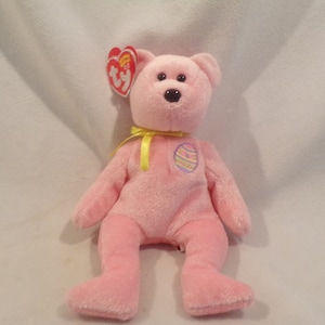 May include: Pink plush Easter bunny with a yellow ribbon and an embroidered Easter egg on its chest. The bunny is sitting on a white surface.