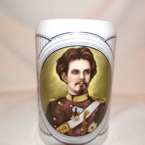 May include: A white ceramic beer stein with a portrait of a man in a military uniform. The portrait is surrounded by a gold and white decorative border.