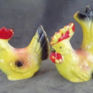 May include: A pair of ceramic rooster salt and pepper shakers. The rooster on the left is yellow with a red comb and black tail feathers. The rooster on the right is yellow with a red comb and black tail feathers.
