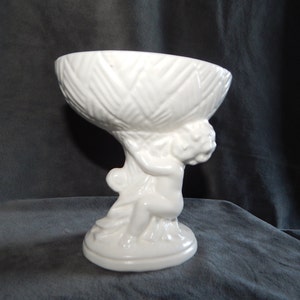 May include: White ceramic bowl with a textured design, held by a cherub figurine. The bowl is on a pedestal base.