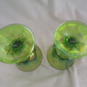 May include: Two green glass cups with a iridescent finish. The cups have a wide base and a narrow top. The base of the cups have a decorative design.