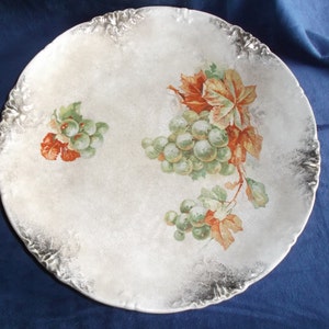 May include: A white porcelain plate with a decorative gold rim and a painted design of green grapes and red leaves.
