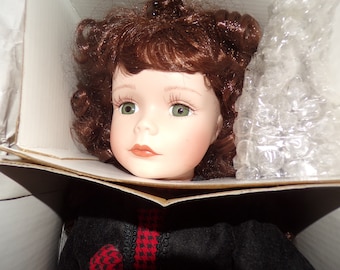 May include: A porcelain doll with brown curly hair and green eyes, wearing a black dress with a red and black plaid accent. The doll is in a box with packing peanuts.