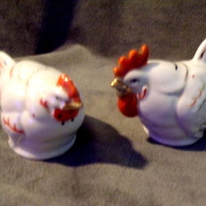 May include: A pair of ceramic rooster and hen figurines. The white figurines have red combs and wattles, with gold beaks and accents. The hen is on the left, and the rooster is on the right. The figurines are likely salt and pepper shakers.