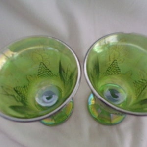 May include: Two green glass goblets with a grape design. The goblets have a silver rim and a iridescent finish.