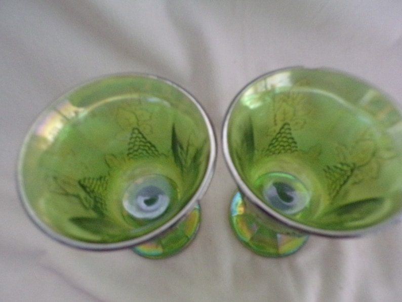 May include: Two green glass goblets with a grape design. The goblets have a silver rim and a iridescent finish.