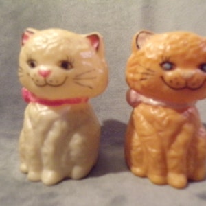 May include: Two ceramic cat figurines, one white and one orange, with pink bows around their necks. They are both sitting with their paws tucked under them and have painted facial features.