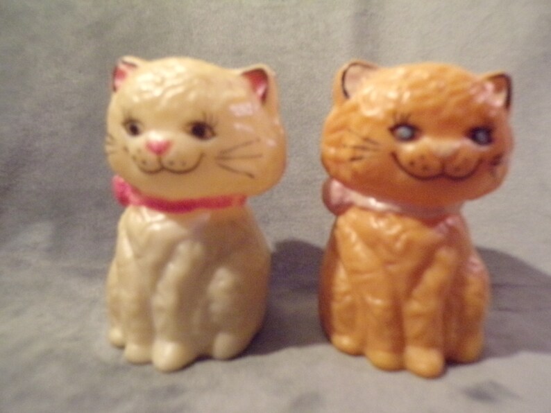 May include: Two ceramic cat figurines, one white and one orange, with pink bows around their necks. They are both sitting with their paws tucked under them and have painted facial features.
