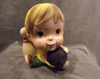 May include: A ceramic figurine of a child holding a bowling ball and a bowling pin. The child has blonde hair, blue eyes, and freckles. The figurine is painted in bright colors.