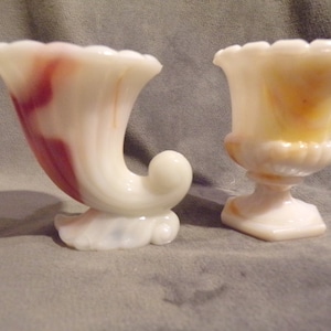 May include: Two antique glass vases. One is a white cornucopia shape with red and white swirls. The other is a white urn shape with yellow and white swirls. Both have scalloped rims and sit on a white base.
