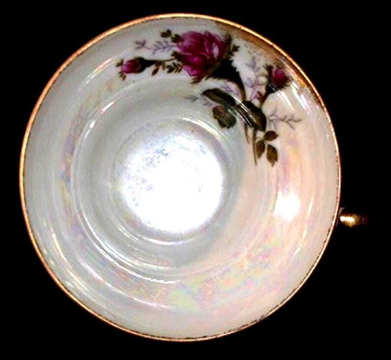 May include: A round porcelain teacup with an iridescent finish and a gold-trimmed rim. The interior features a floral motif with a single large magenta rose and two smaller rosebuds in shades of pink and green, set against a white background.