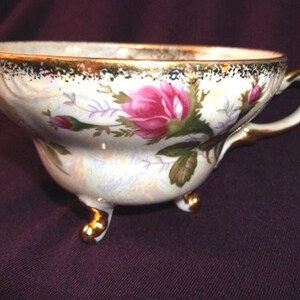 May include: A porcelain teacup featuring an iridescent finish, decorated with pink rose blossoms and green foliage. The rim and handle are accented with gold trim, and the cup is supported by three small, gold-painted feet.