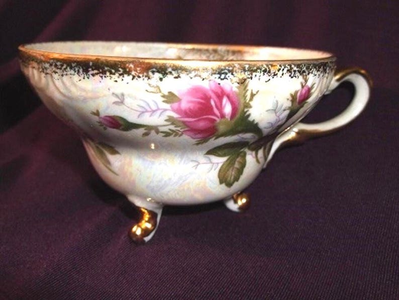 May include: A porcelain teacup featuring an iridescent finish, decorated with pink rose blossoms and green foliage. The rim and handle are accented with gold trim, and the cup is supported by three small, gold-painted feet.