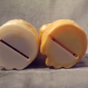 May include: Two plastic piggy banks, one white and one tan, with a slot on the bottom for coins.