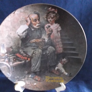 May include: A vintage porcelain plate featuring a painting of a man with a worried expression, a young girl holding a flower, and a child lying on the steps. The plate has a brown background and the artist's signature is visible in the bottom right corner.