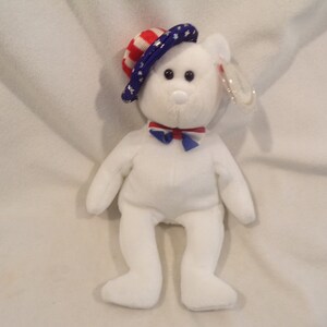 May include: A white plush bear wearing a patriotic hat with red and white stripes and blue stars. It has a red and blue bow tie. The bear is a stuffed animal, likely a collectible item.
