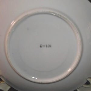 May include: Underside of a white porcelain plate with a scalloped edge. The plate has a circular recessed area in the center. The text "C-131" is printed in black near the center. The plate is resting on a gold-colored stand.