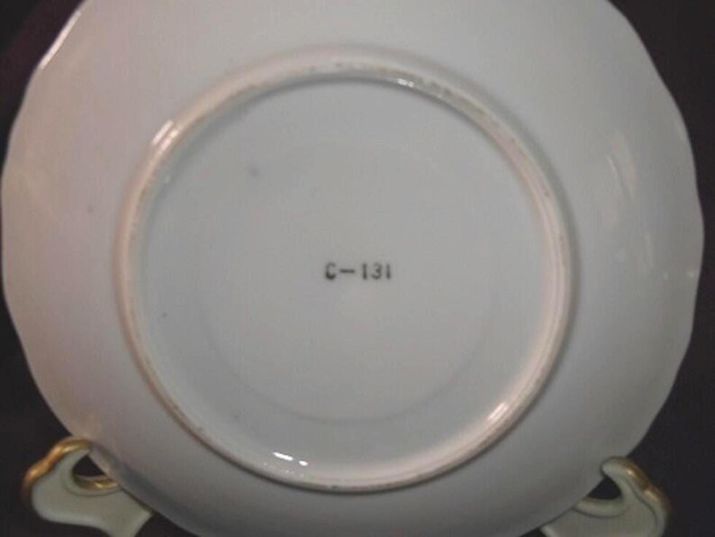 May include: Underside of a white porcelain plate with a scalloped edge. The plate has a circular recessed area in the center. The text "C-131" is printed in black near the center. The plate is resting on a gold-colored stand.