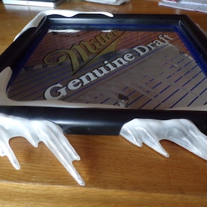 May include: A black framed sign with the Miller Genuine Draft logo. The sign has a clear glass surface and is decorated with white, icicle-shaped accents along the edges. The background is a wooden surface.