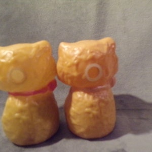 May include: Two yellow ceramic cat figurines with a textured surface. One cat has a red ribbon around its neck.