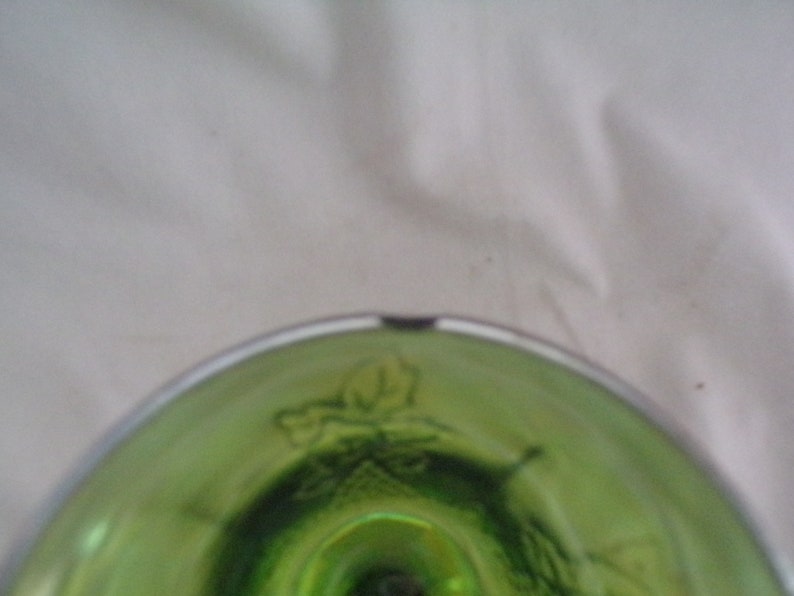 May include: Close-up of the bottom of a green glass goblet with a white rim. The goblet has a decorative design of leaves and vines.