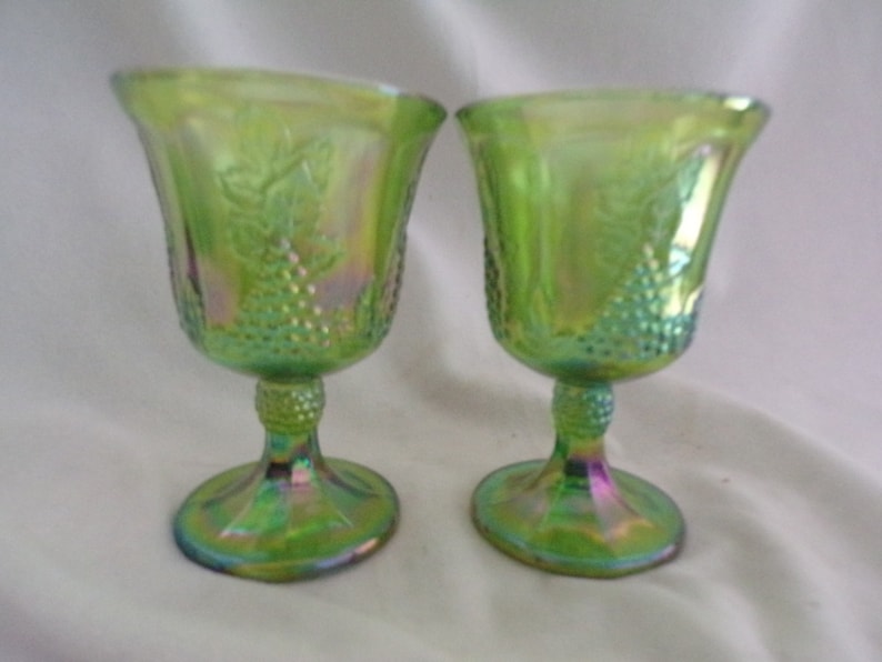 May include: Two green iridescent glass goblets with a grape design. The goblets have a stemmed base and a wide bowl.