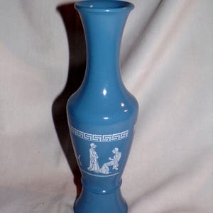 May include: A light blue vase with a flared top and base. The vase features a white Greek key design and white figures. The vase is approximately 9 inches tall.