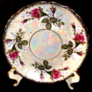 May include: A round porcelain saucer featuring an iridescent finish and a scalloped edge with a gold-tone trim. The surface is decorated with clusters of pink rose blossoms, green leaves, and small buds arranged around a central circular area.