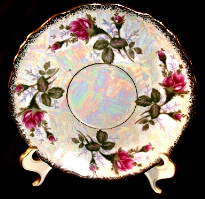 May include: A round porcelain saucer featuring an iridescent finish and a scalloped edge with a gold-tone trim. The surface is decorated with clusters of pink rose blossoms, green leaves, and small buds arranged around a central circular area.