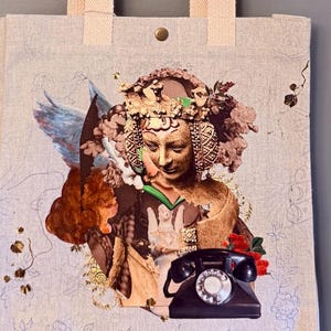 Könnte beinhalten: Eine hellbeige Tragetasche aus Segeltuch mit zwei passenden Stoffgriffen und einem einzelnen Metalldruckknopf. Die Vorderseite zeigt einen Collage-Druck einer goldfarbenen Figur mit einem verzierten Kopfschmuck, einem schwarzen Wählscheibentelefon.