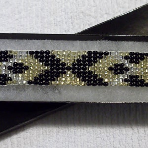 May include: A black and gold beaded bracelet with a geometric pattern. The bracelet is in a black box with white lining.