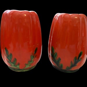 May include: Two red ceramic tulip-shaped vases with a glossy finish. Each vase has a scalloped rim and green leaf design near the base. The vases are set against a black background.