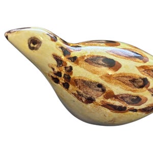 May include: A ceramic bird figurine with a light brown and tan speckled glaze. The bird has a rounded body and a long, thin beak.